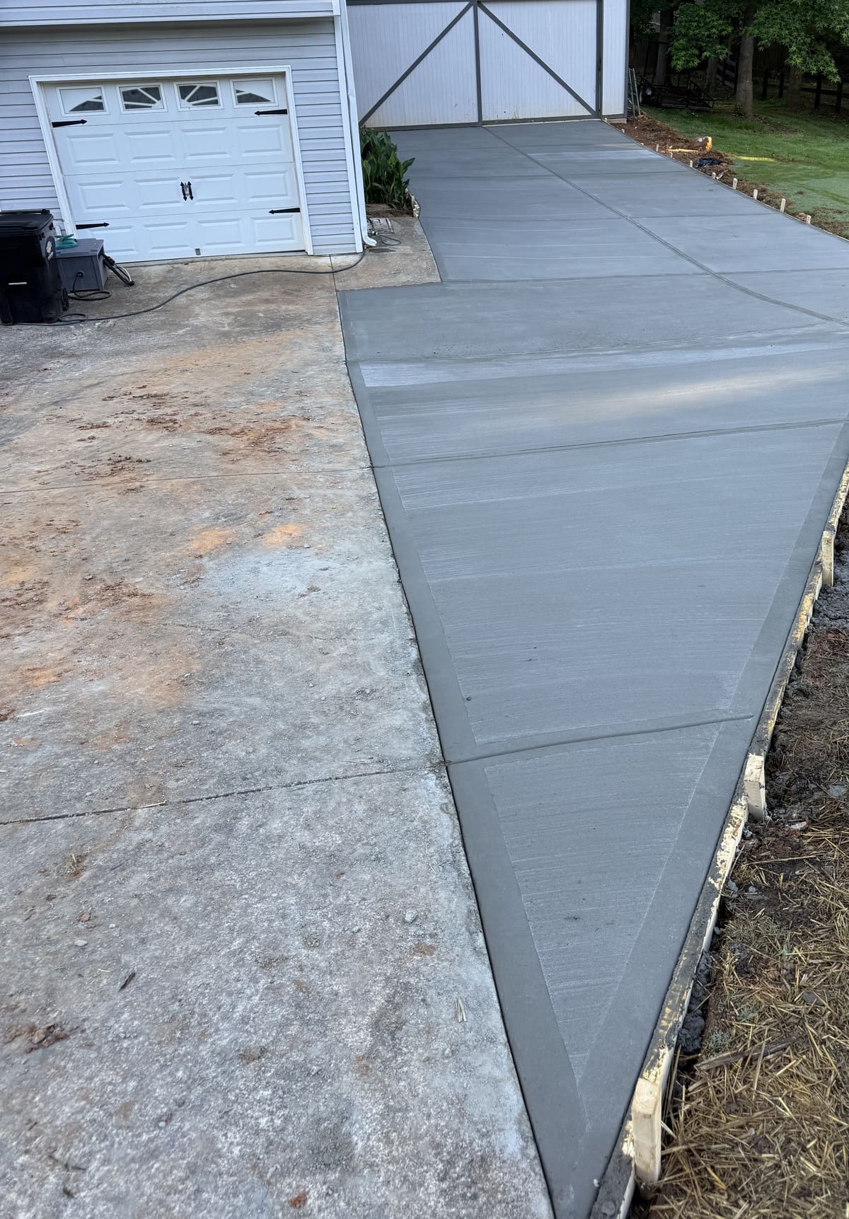 Broom-finished concrete strip widening an older driveway, running past a gray house to a white metal garage