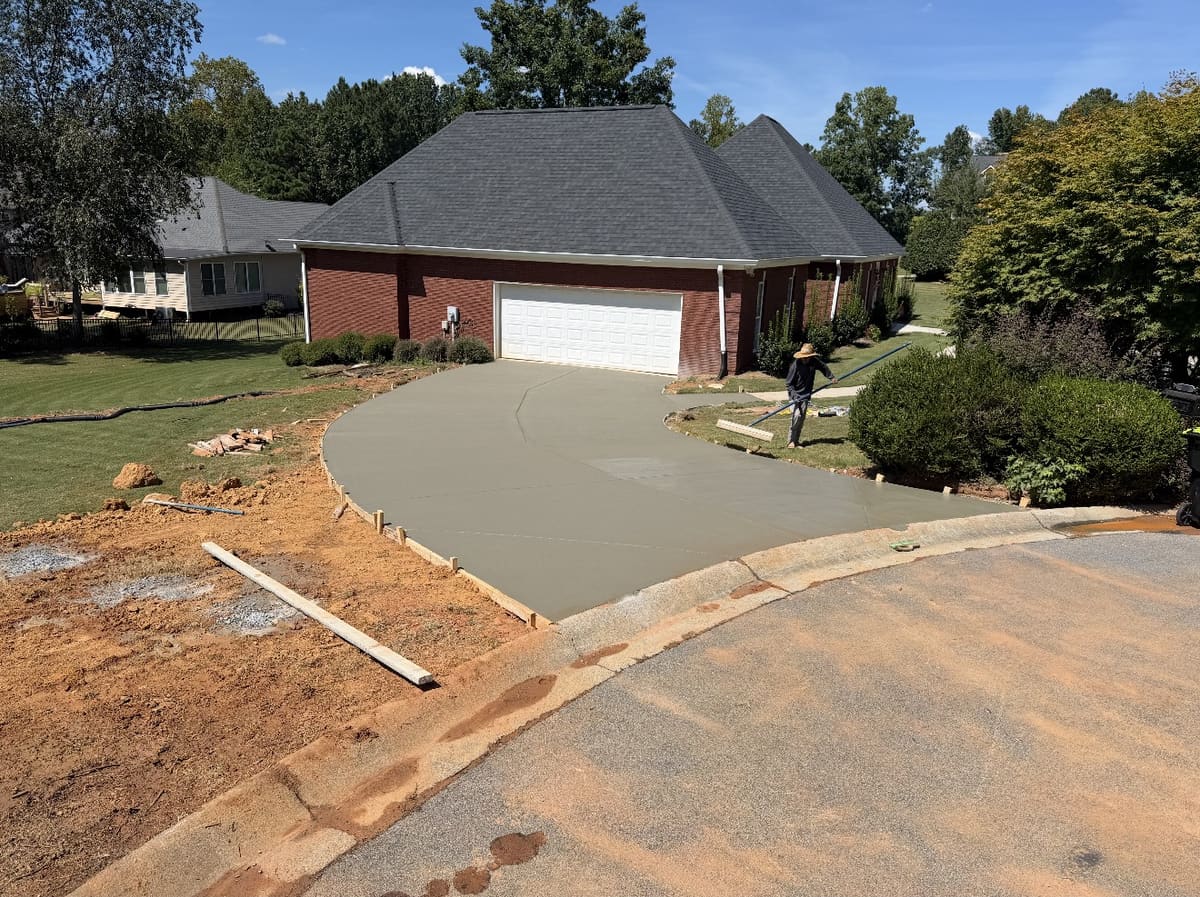 Fresh driveway curving from the street up to a two-car garage on a red brick house, a crew member working a bull float
