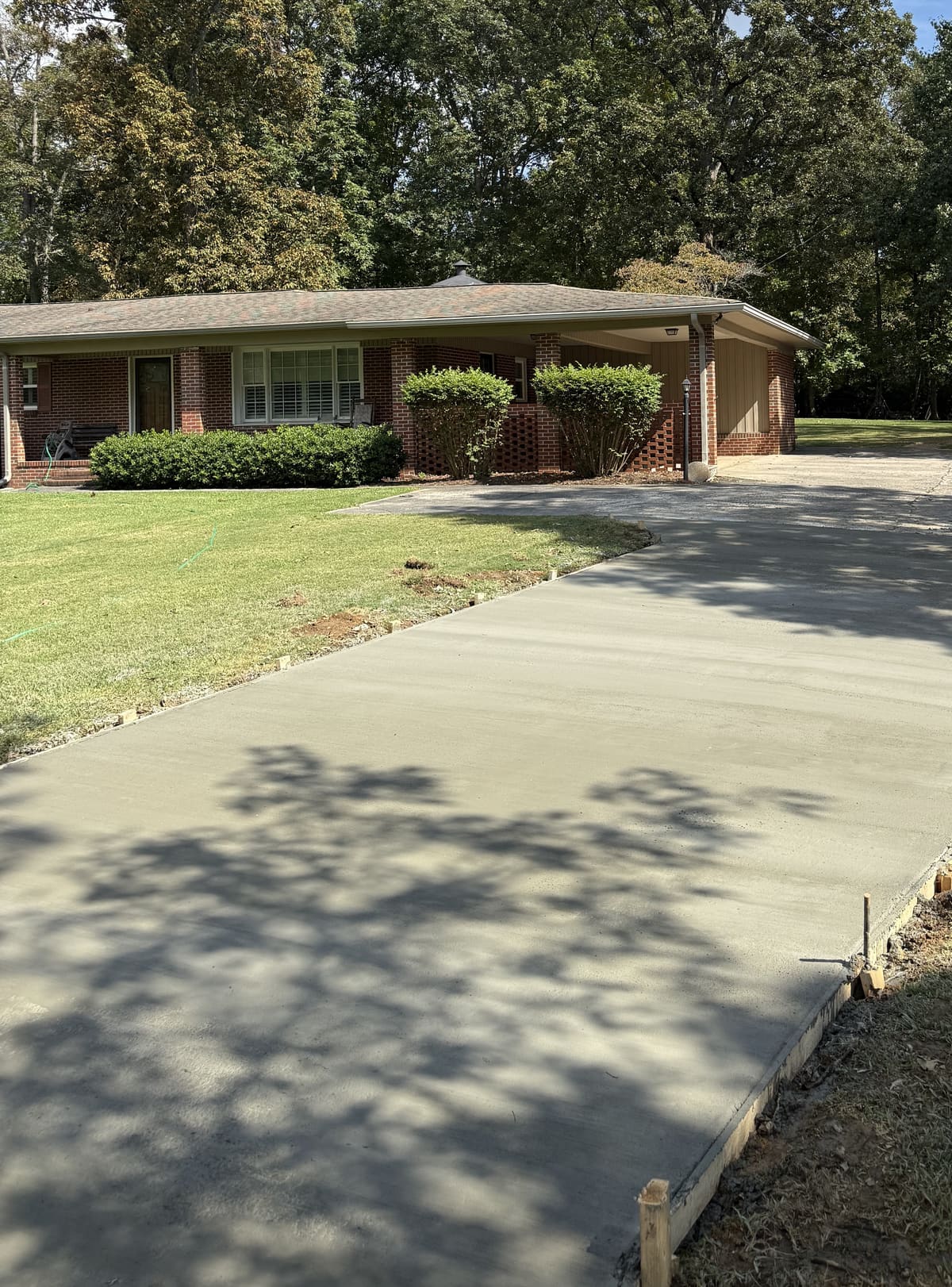 Fresh broom-finished driveway running past a brick ranch house with a carport, tall oaks overhead