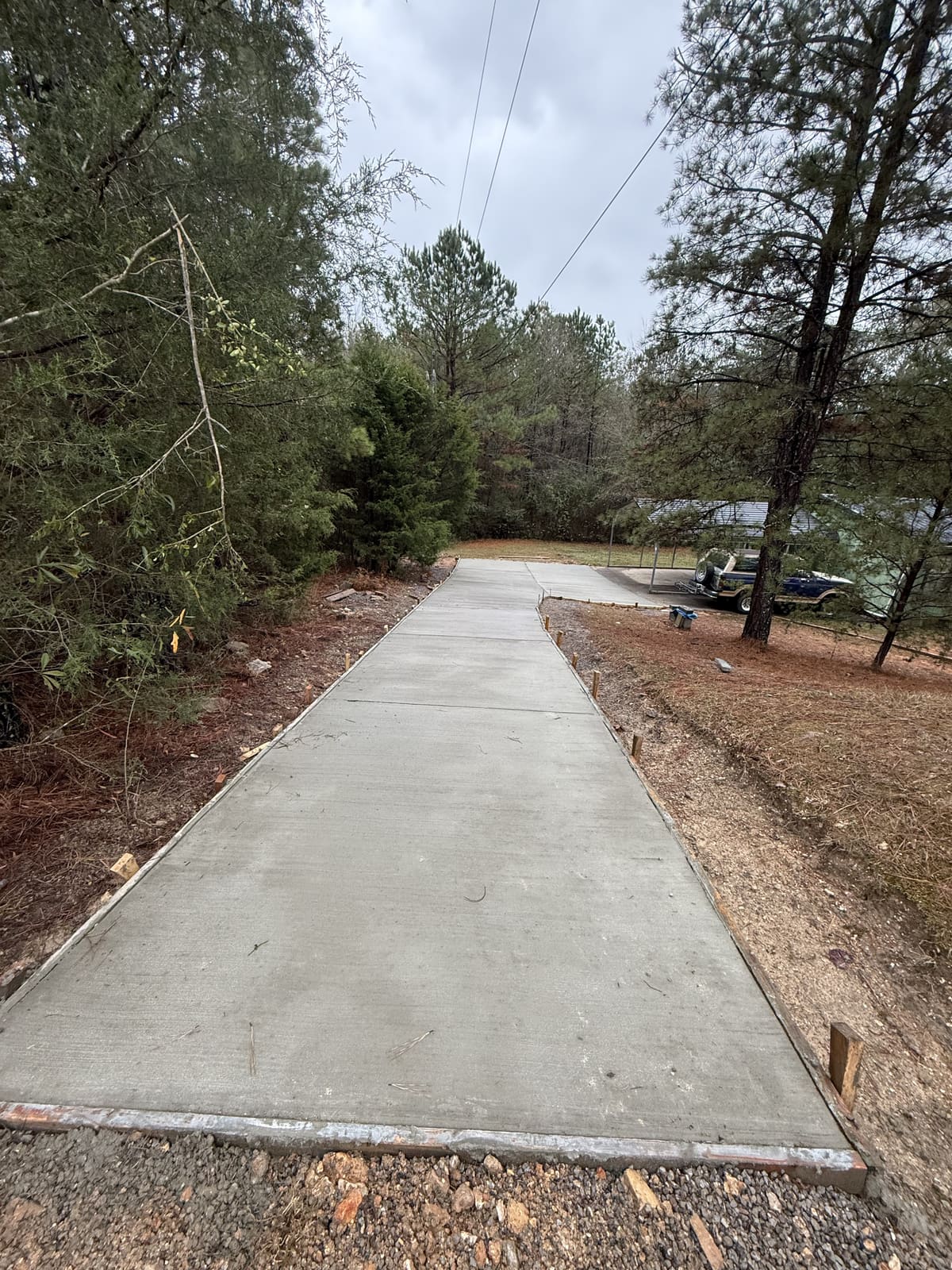 Fresh broom-finished driveway with control joints running downhill through pine woods to a carport
