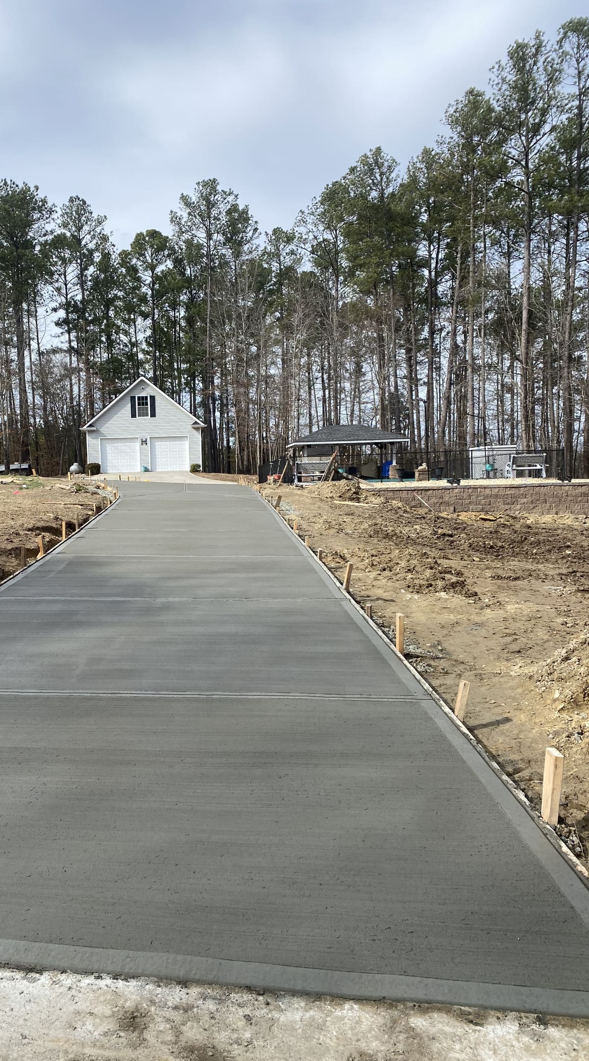 Fresh broom-finished driveway with control joints running between wood forms to a detached two-car garage in the pines