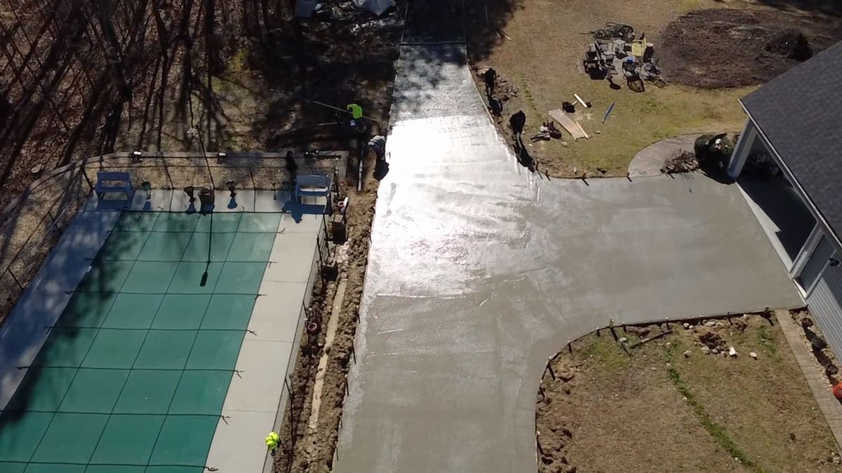 Overhead view of a freshly poured driveway, still wet, running past a covered pool with crew finishing its edges