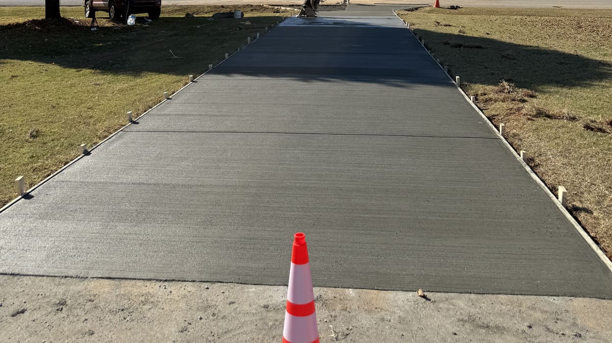 Fresh broom-finished driveway between wood forms with a traffic cone at the near end, crew finishing the far end