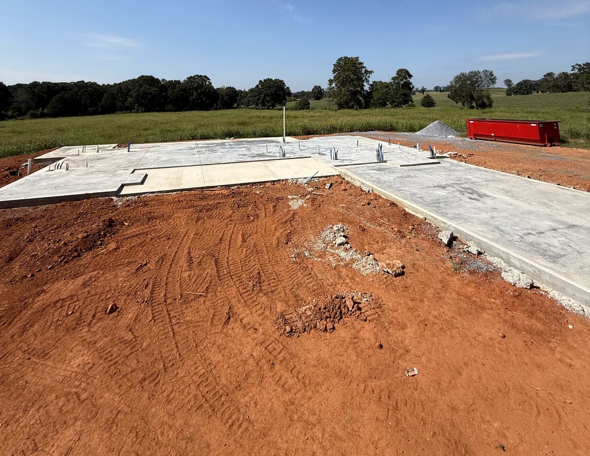Cured house slab with plumbing stubs and the forms stripped, red clay graded around it and open pasture beyond