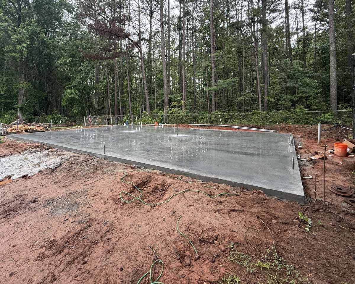 Freshly finished pad with anchor bolts standing along its edges, the surface still wet, tall pines behind