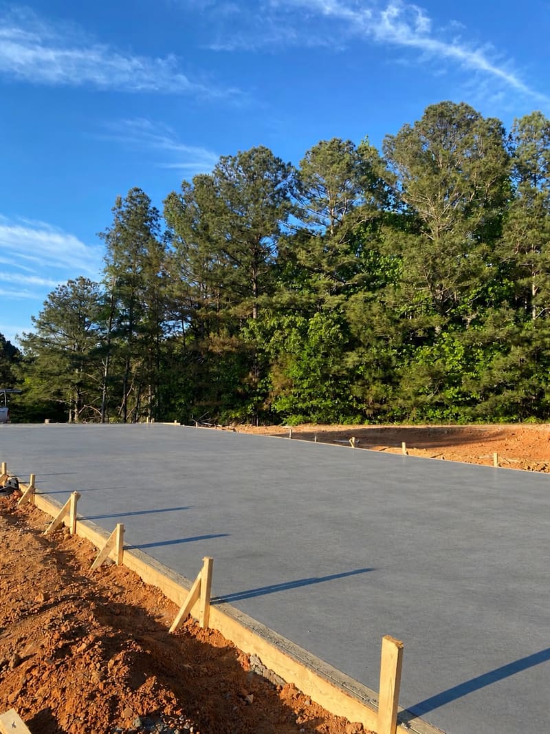 Large fresh pad with a smooth troweled finish inside braced wood forms, pine trees under a blue sky