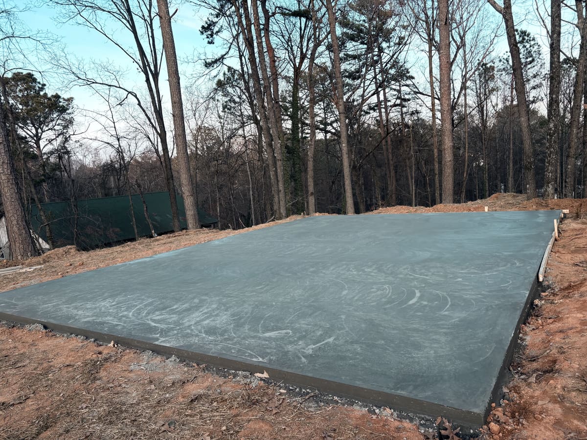 Smooth-troweled pad on a cleared wooded hillside, a green metal roof visible through the trees