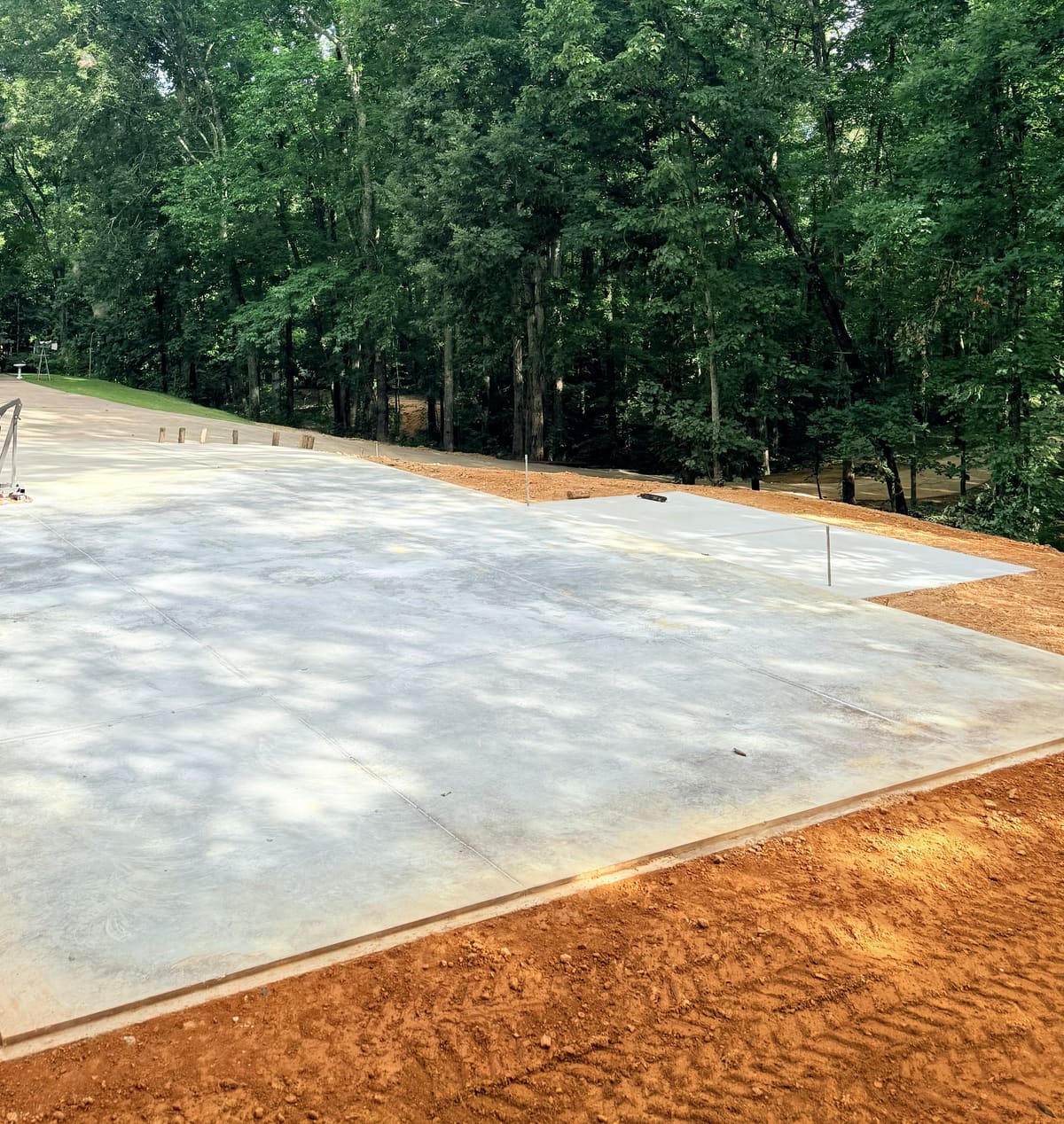 Large cured pad with control joints below tall hardwoods, a small raised section at its back corner