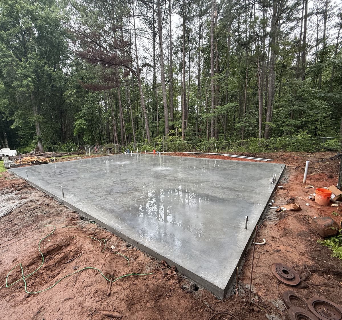 Fresh pad with anchor bolts set along the edges and puddles on its wet surface, pine woods behind