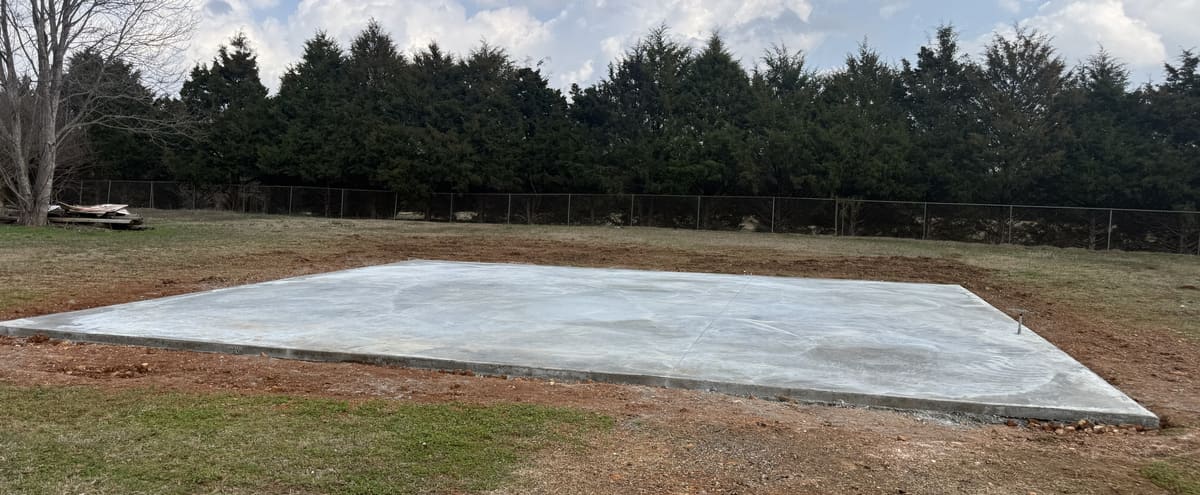 Cured pad on a graded lot in front of a row of cedar trees and a chain-link fence
