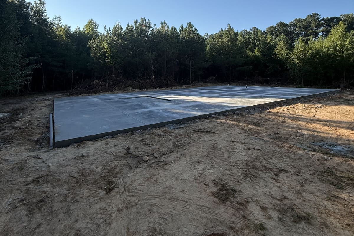 Cured slab with plumbing stubs and a grid of control joints at the edge of young pine woods