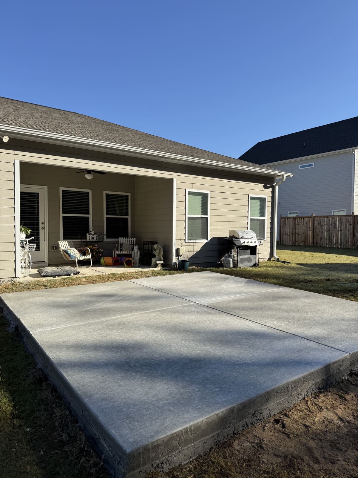 New broom-finished patio slab in four panels behind a tan house with a covered porch and a grill