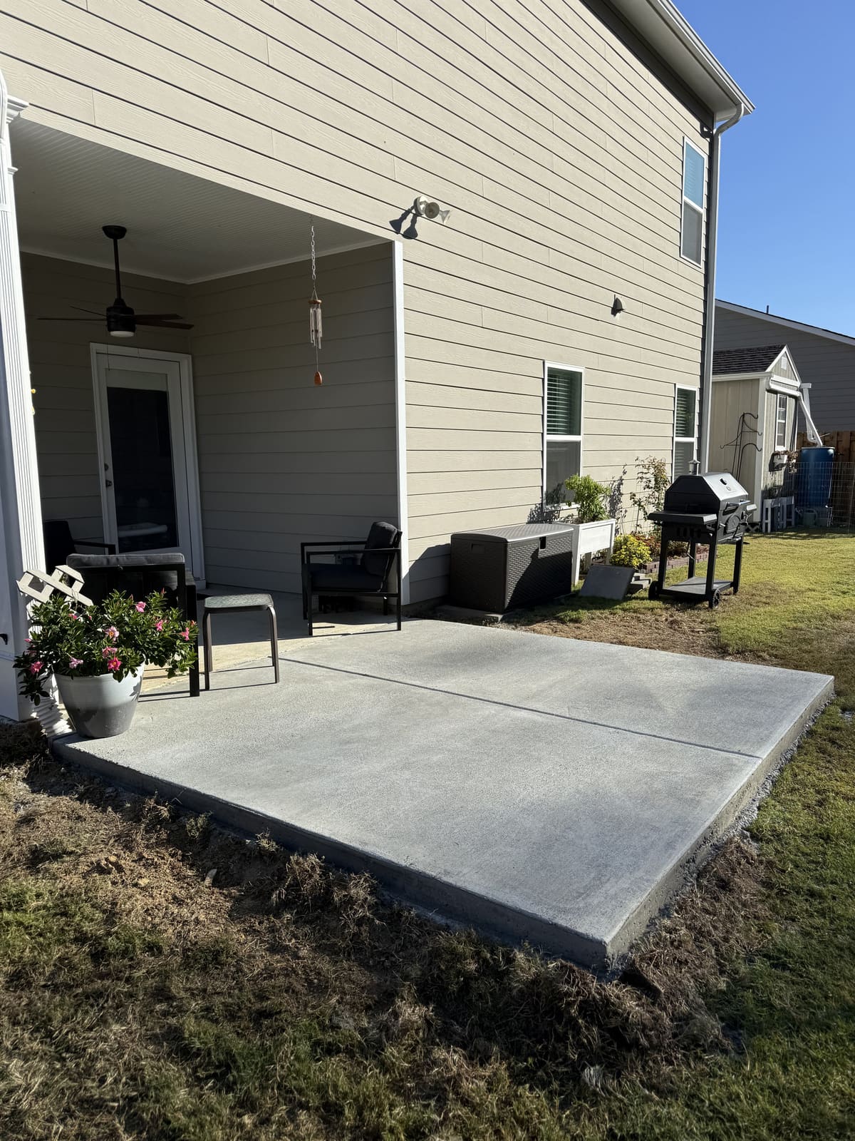New patio slab extending a covered back porch beside a tan two-story house, a grill at its far corner