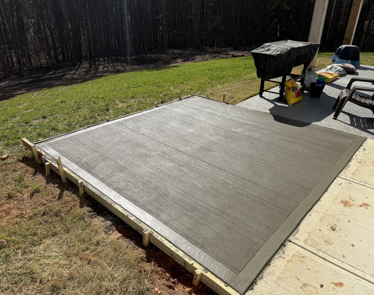 Fresh broom-finished patio extension in wood forms beside a covered porch, a grill and lawn chair at its edge