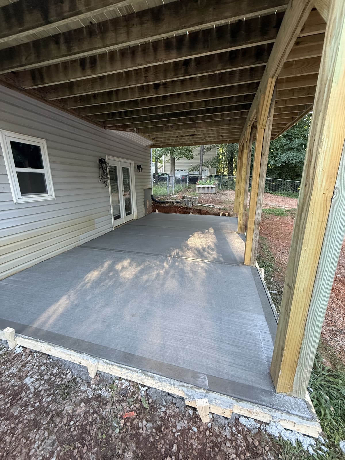 Fresh broom-finished patio poured under a raised wood deck beside a gray house with French doors
