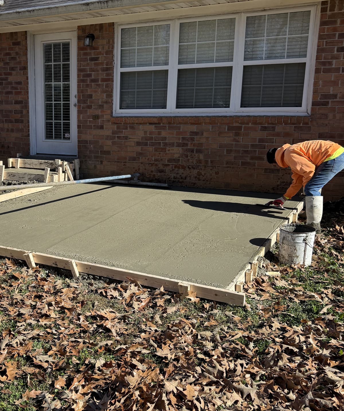 Crew member edging a freshly placed slab in wood forms against a brick house, a formed step at the door beside it