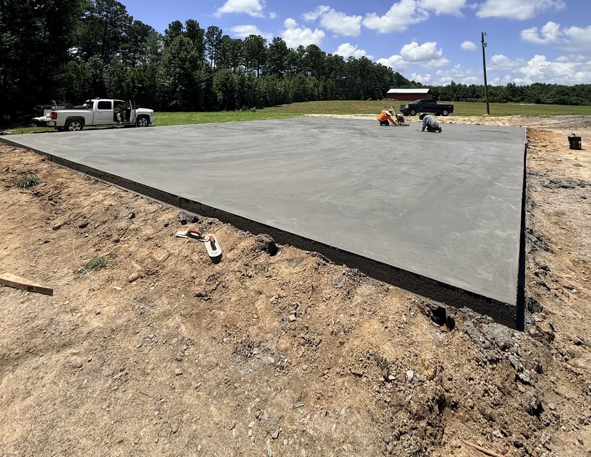 Crew finishing a large fresh pad with a power trowel and hand tools in open pasture, a red barn far off