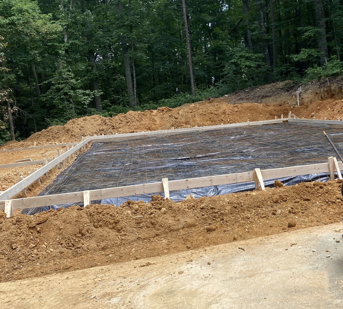 Black vapor barrier and wire mesh inside wood forms, ready for a pour below a wooded slope