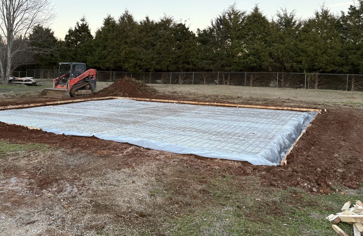 Vapor barrier and wire mesh inside wood forms, a tracked skid steer parked by the dirt pile behind