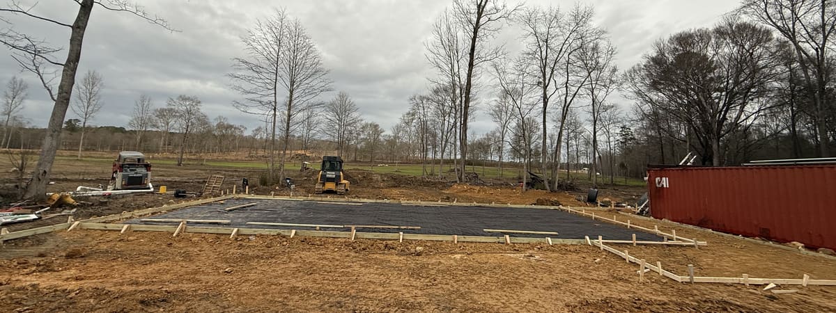 Black vapor barrier and wire mesh in long wood forms, a dozer and skid steer on the graded lot behind