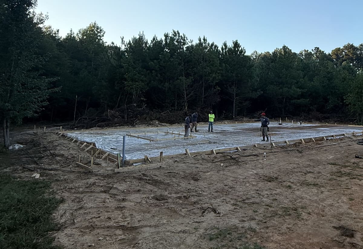 Crew walking wire mesh over vapor barrier inside large wood forms, a plumbing riser in front, pine woods behind
