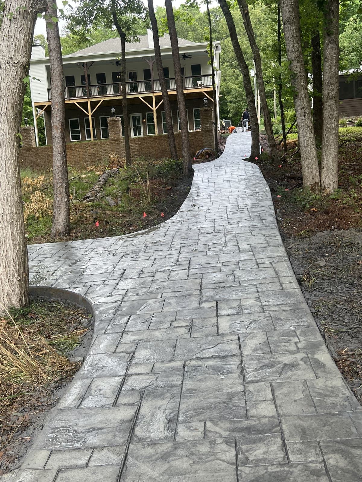 Gray stamped walkway in an ashlar stone pattern winding uphill through the trees to a large two-story house