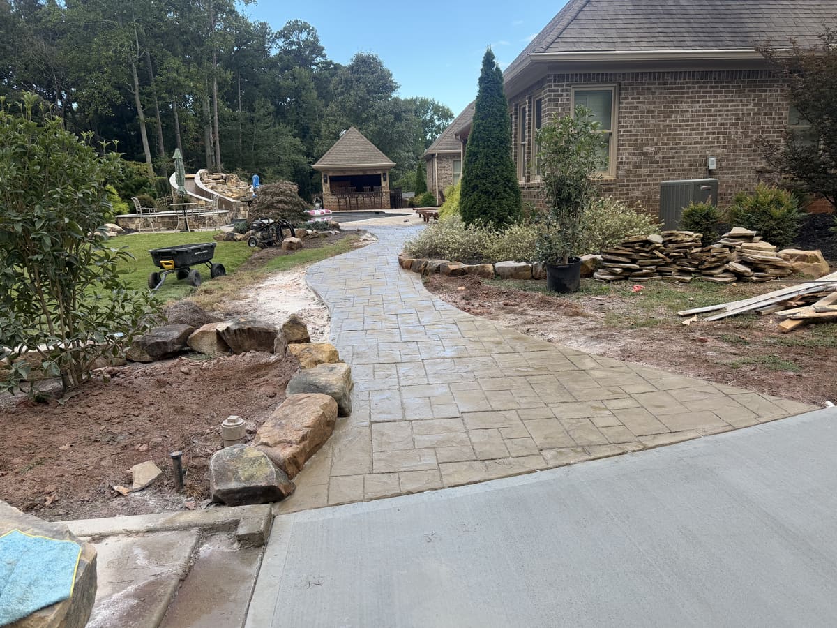 Tan stamped walkway in an ashlar stone pattern curving past a brick house toward a pool house, boulders along one side
